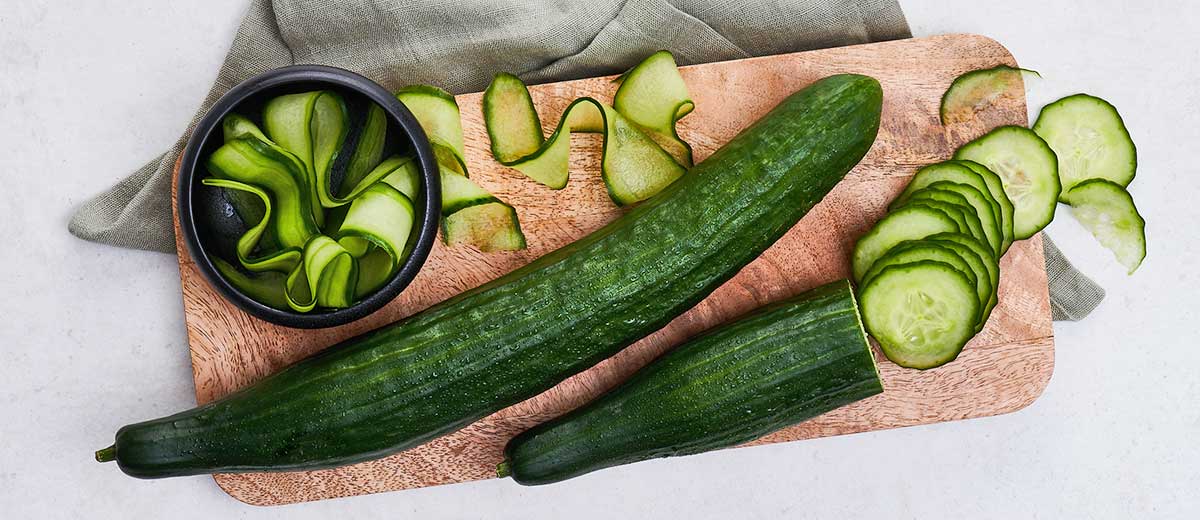 Abbildung: Frische Gurke im Ganzen, geschnitten und gehobelt auf einem Holzschneidebrett angerichtet