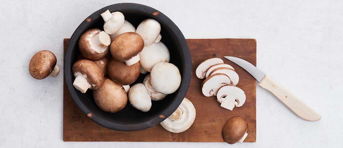 Abbildung: Champignons in einer Schüssel, die auf einem Holzbrett steht. Daneben liegt ein Messer